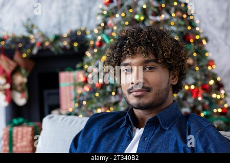 Closeup portrait of a young serious man, hispanic sitting at home on the couch and looking thoughtfully and focused at the camera, celebrating the new year and Christmas, near tree decorated gifts. Stock Photo