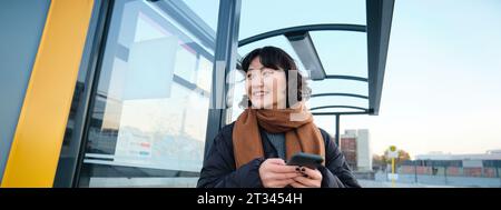 Cute smiling asian girl standing on bus stop, holding smartphone, wearing winter jacket and scarf. Woman commuting to work or university via public Stock Photo