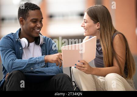 Young beautiful couple smiling and reading together a notebook.Two diverse students sitting outside talking while holding files. Stock Photo