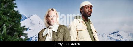 stylish multiethnic couple in fashionable jackets with mountain backdrop, winter fashion, banner Stock Photo