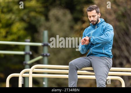 A young athlete sits on the bars in nature and monitors his activity on a smart watch. Stock Photo