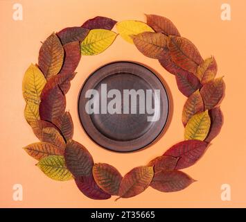 Wreath from Colorful fall leaves and empty brown plate inside it on pastel orange background. Autumn, Halloween, thanksgiving day mock up. Top view. F Stock Photo