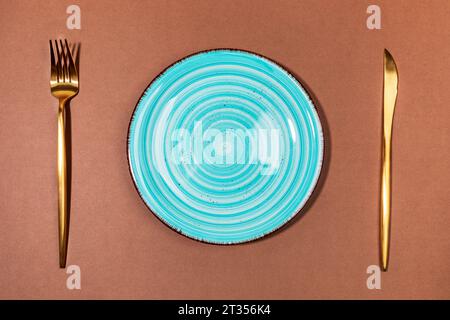One round empty turquoise tableware plate and golden cutlery knife and fork on brown backdrop. Idea fall holiday table setting for Halloween or thanks Stock Photo