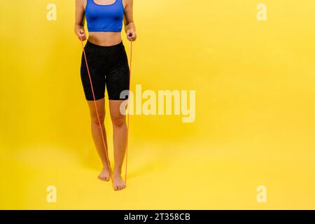 Fit female athlete with ponytail jumping with rope during fitness training against yellow background Stock Photo