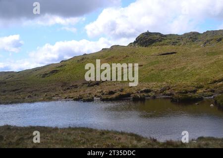 The Trig Point on the Summit of the Wainwright on Place Fell from a  Small Tarn near Hart Crag, Lake District National Park, Cumbria, England, UK. Stock Photo