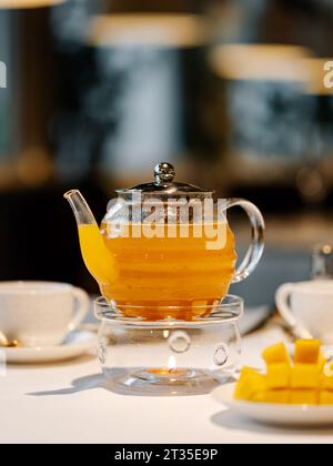 Herbal tea with mango fruit in transparent glass teapot with warmer and candle on table in restaurant. Herbal tea concept, wellbeing and health care. Vertical. Stock Photo