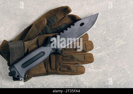 Military tactical knife lying on gloves, gray concrete background Stock Photo