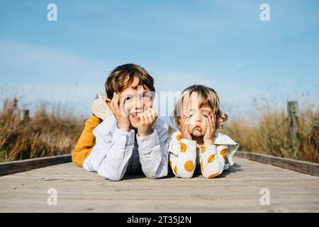 Portrait of boy and his little sister lying side by side on boardwalk pulling funny faces Stock Photo