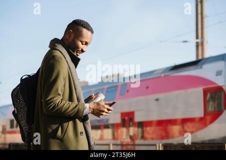 Stylish man with reusable cup using smartphone while waiting for the train Stock Photo