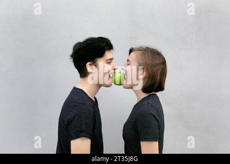 Teenage couple carrying apple in mouth together against gray background Stock Photo