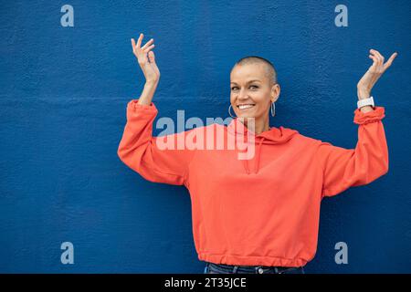 Smiling woman with shaved head dancing in front of blue wall Stock Photo