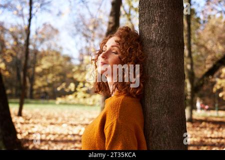 Redhead woman with eyes closed leaning against tree at autumn park Stock Photo