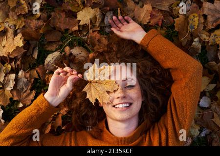 Smiling redhead woman holding maple leaf and lying on ground at park Stock Photo