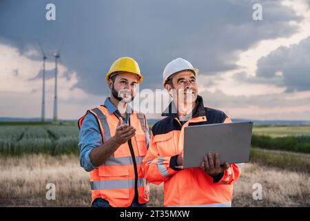 Smiling engineers working with laptop at wind farm Stock Photo