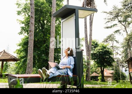 Woman sitting with laptop near solar charging point at park Stock Photo