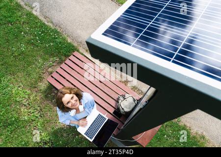 Smiling woman with laptop looking at solar charging point Stock Photo
