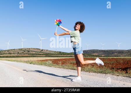 Happy woman standing with multi colored pinwheel toy in front of wind turbines Stock Photo