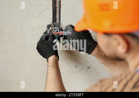 Electrician male in uniform, protective gloves and helmet checks presence of electrical voltage in socket phase uses electrical tester screwdriver.Exa Stock Photo