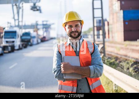 Smiling logistcs worker wearing hardhat holding tablet PC standing on road Stock Photo