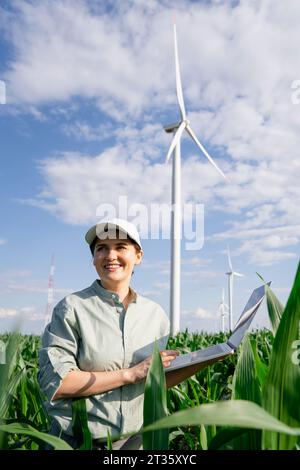 Happy agronomist standing with laptop in front of wind turbines on sunny day Stock Photo
