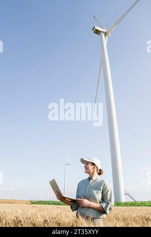 Happy farmer with laptop standing in front of wind turbines on sunny day Stock Photo