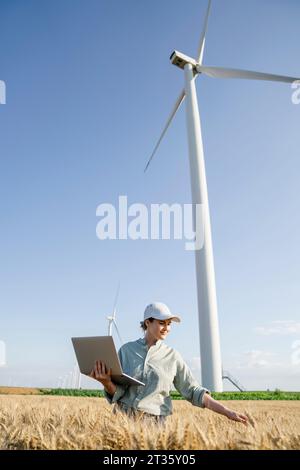 Smiling farmer with laptop examining crops in front of wind turbines on sunny day Stock Photo