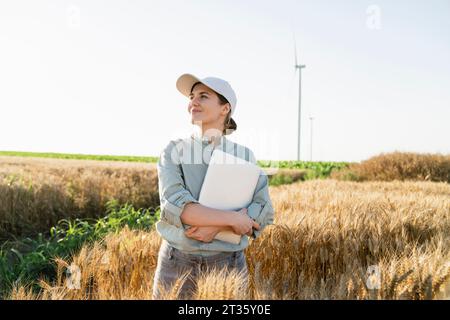 Smiling farmer wearing cap and holding laptop at wheat field Stock Photo