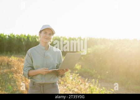 Smiling agronomist standing with laptop in field Stock Photo