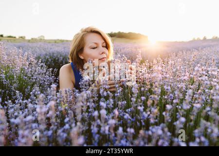 Smiling woman smelling lavender flowers in field at sunset Stock Photo