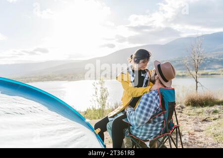 Girlfriend sitting on boyfriend's lap by tent near lake Stock Photo