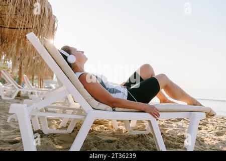 Woman wearing headphones relaxing on lounge chair at beach Stock Photo