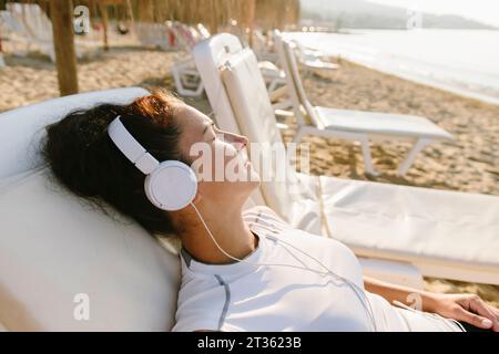 Woman wearing headphones resting on lounge chair at beach Stock Photo