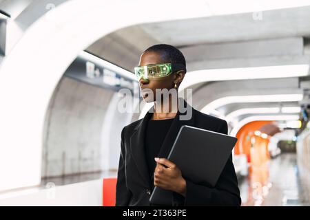 Contemplative young woman wearing smart glasses and standing with laptop at metro station Stock Photo