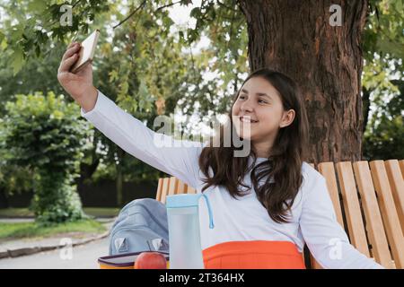 Smiling schoolgirl taking selfie with smart phone sitting on bench Stock Photo