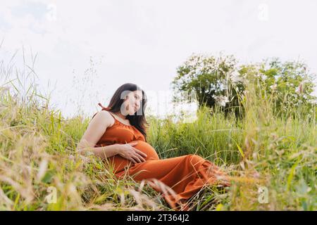 Pregnant woman sitting on grass under sky Stock Photo