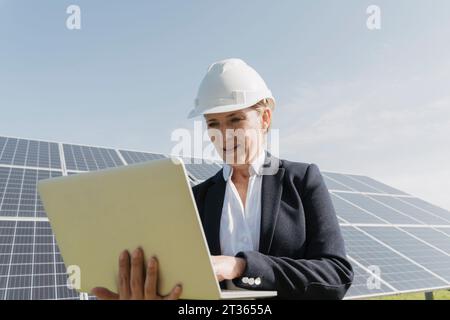 Engineer wearing hardhat working on laptop in front of solar panels Stock Photo