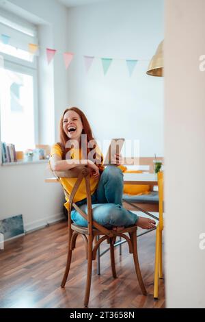 Cheerful woman laughing and sitting with tablet PC on chair at home Stock Photo