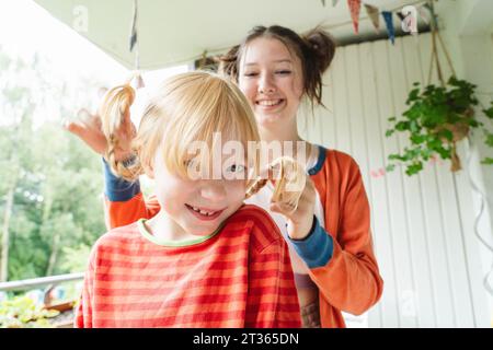 Blond girl being braided by her happy teenage sister on balcony Stock Photo