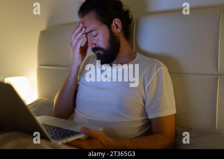 Man with laptop having headache on bed at home Stock Photo