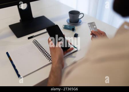 Man holding smart phone and blister pack at table Stock Photo