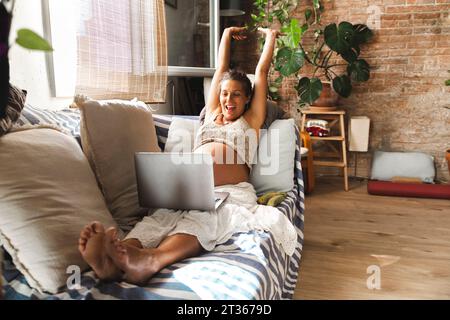 Smiling pregnant woman sitting with arms raised and laptop on sofa Stock Photo