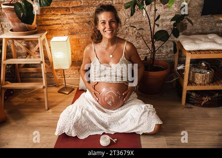 Smiling pregnant woman applying moisturizer on abdomen at home Stock Photo