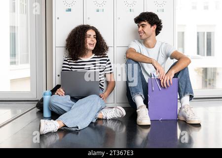 Smiling students with laptop sitting in front of locker in university Stock Photo