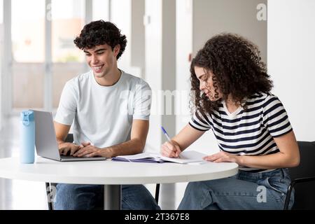 Smiling students writing on book and using laptop at table in university Stock Photo
