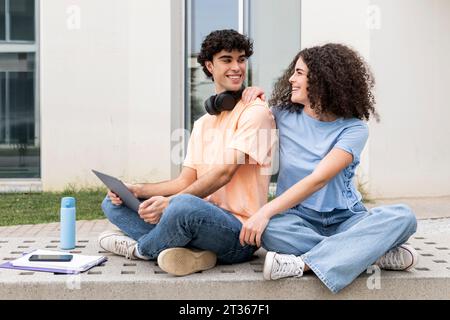 Smiling students sitting with laptop on concrete bench in campus Stock Photo