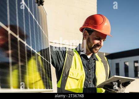 Smiling engineer wearing protective workwear using digital tablet by solar panel Stock Photo