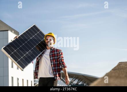 Engineer wearing hardhat carrying solar panel on shoulder at terrace Stock Photo