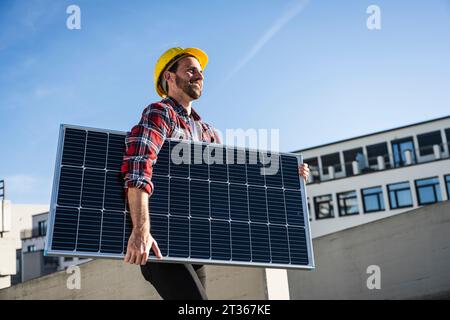 Smiling engineer wearing hardhat carrying solar panel Stock Photo