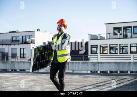 Engineer wearing hardhat carrying solar panel at terrace on sunny day Stock Photo