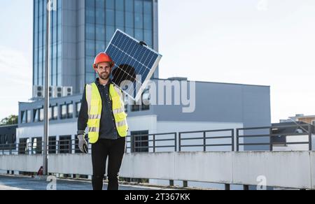 Engineer wearing hardhat carrying solar panel on shoulder at terrace Stock Photo